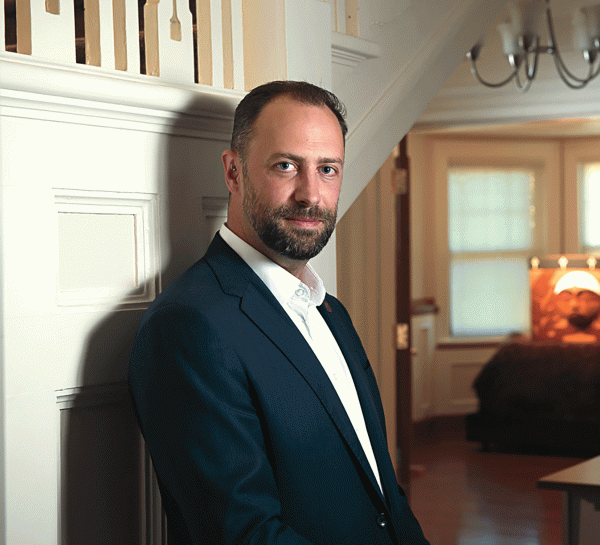 man in a blue suit leaning against a wall looking at a camera