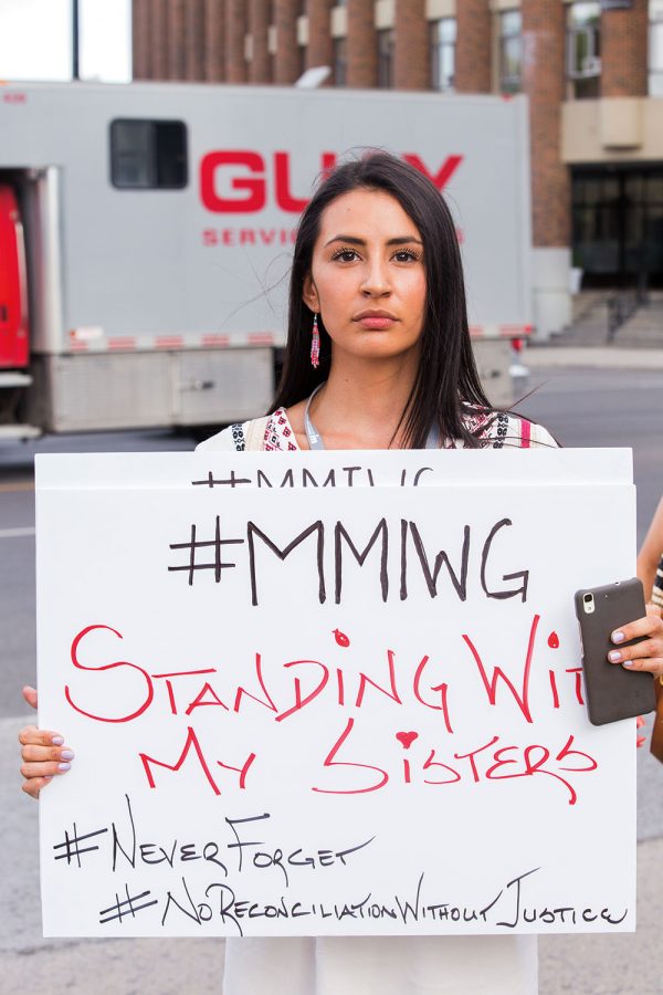 Cree woman holding banner that bears #MMIWG - Standing with my sisters #NeverForger #NoReconcilationWithoutJustice text