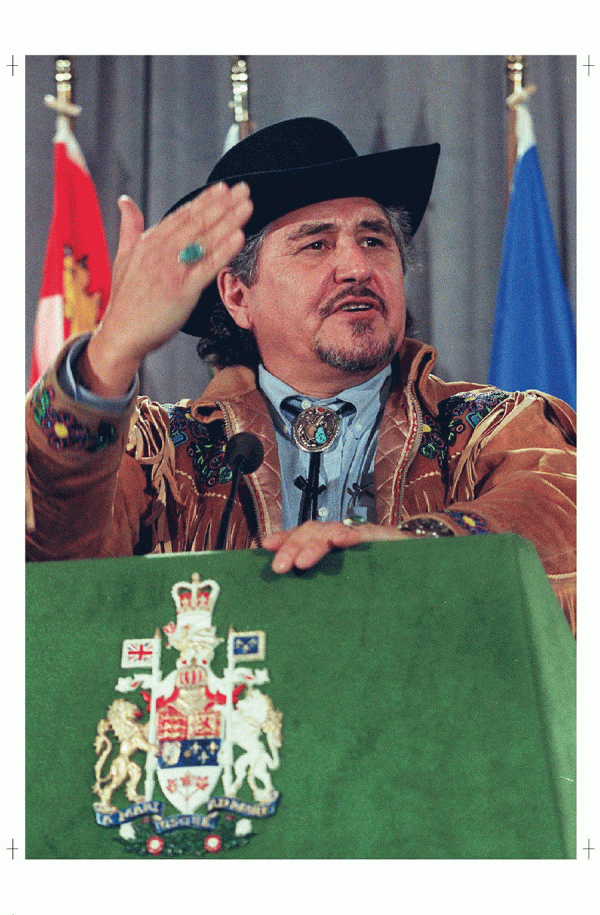 man standing behind a podium wearing brown jacket, black hat, and blue ring