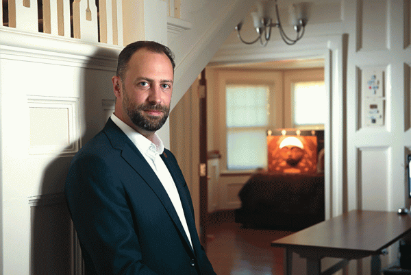 man in a blue suit leaning against a white wall looking at a camera