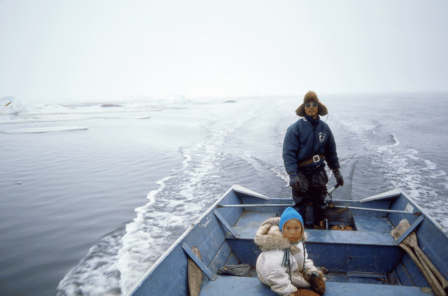 A father and son drive a boat while wearing parkas