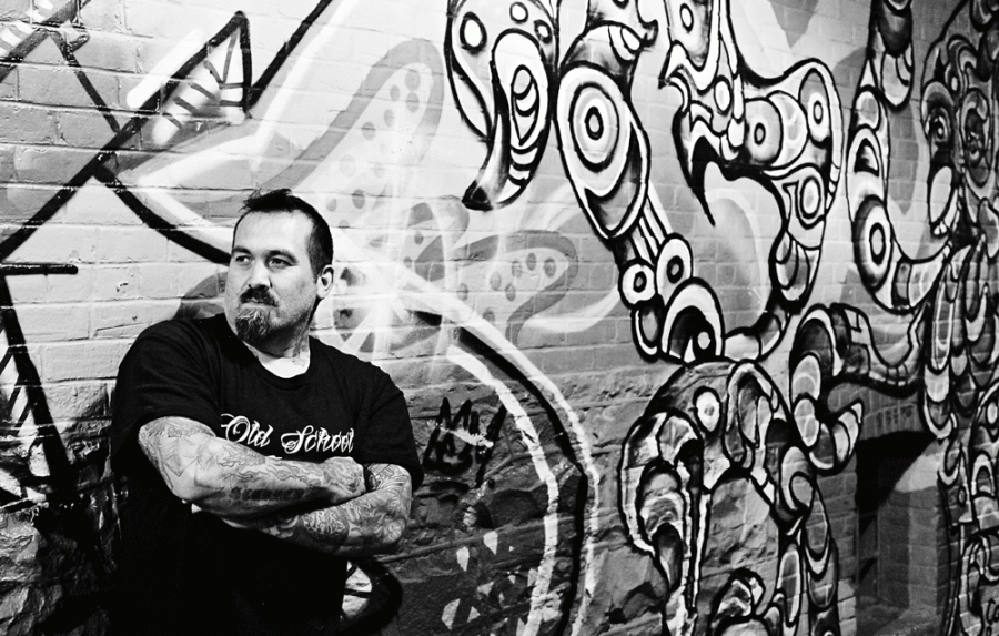 Black and white photo of a man sdtanding with his arms crossed in front of a wall with graffiti on it
